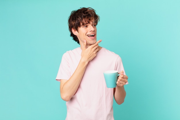 Młody mężczyzna z kubkiem kawy uśmiechający się ze szczęśliwym, pewnym siebie wyrazem twarzy z ręką na brodzie