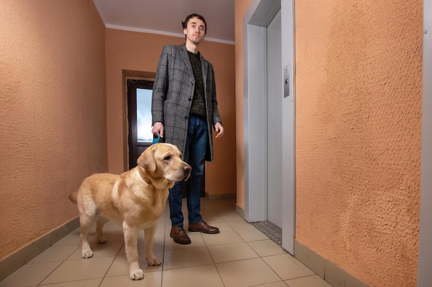 Młody mężczyzna z psem labrador w płaszczu stojący w korytarzu i czekająca na windę