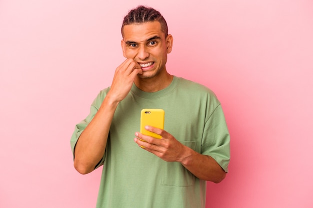 Młody mężczyzna z Wenezueli trzymający telefon komórkowy na białym tle na różowym tle gryzie paznokcie, nerwowy i bardzo niespokojny.