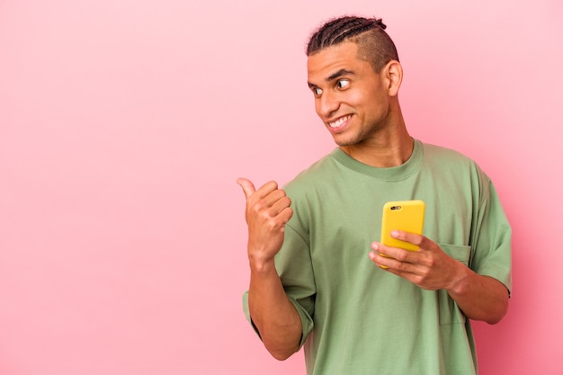 Młody mężczyzna z Wenezueli trzymający telefon komórkowy na białym tle na różowym tle wskazuje palcem kciuka, śmiejąc się i beztrosko.
