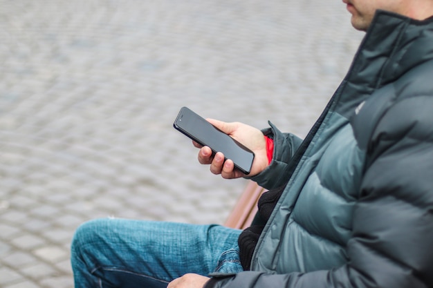 Młody miastowy mężczyzna używa smartphone obsiadanie na ławce w miasto ulicie.