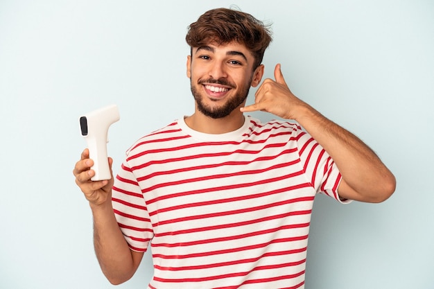 Młody mieszanej rasy mężczyzna posiadający termometr na białym tle na niebieskim tle pokazujący gest połączenia z telefonem komórkowym palcami.