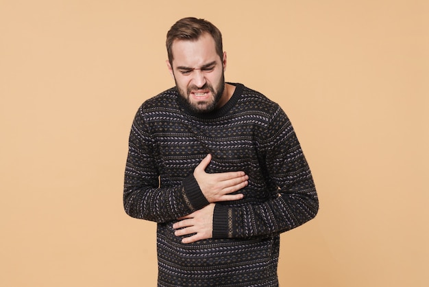 Zdjęcie młody nieogolony mężczyzna w zimowym swetrze dotyka swojego brzucha z powodu bólu izolowanego na beżowej ścianie