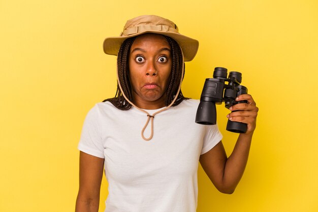 Młody odkrywca Afryki amerykański kobieta trzyma lornetkę na białym tle na żółtym tle wzrusza ramionami i otwiera oczy zdezorientowany.