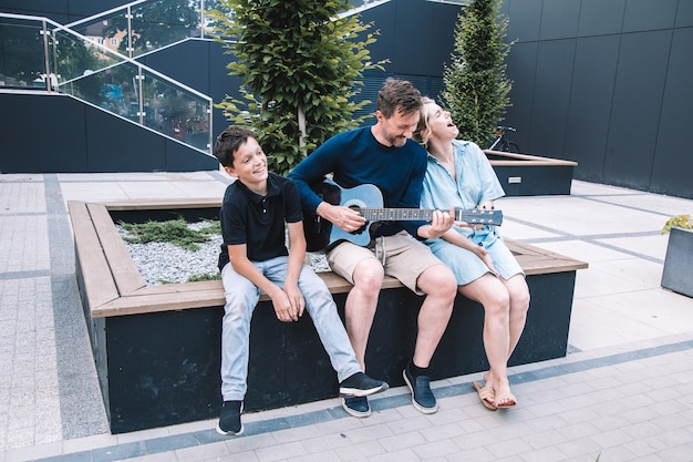 Młody ojciec gra wesołą piosenkę na gitarze dla swojej rodziny w letni dzień na placu miejskim. Styl życia.