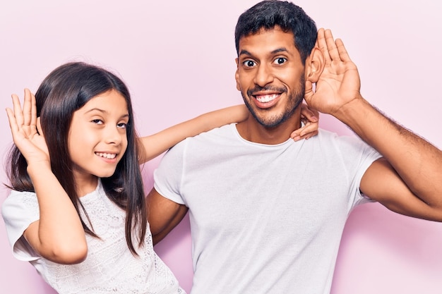 Młody ojciec i córka w przypadkowych ubraniach uśmiechający się z ręką nad uchem słuchający koncepcji głuchoty plotek lub plotek
