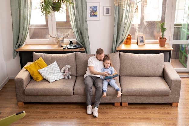 Młody ojciec i jego urocza córeczka siedzą na kanapie i czytają bajki, przebywając w domu na kwarantannę