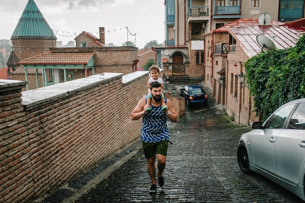Młody ojciec spaceruje z córeczką na świeżym powietrzu po ulicach starego miasta Tbilisi w stolicy Gruzji w deszczowy dzień Tata i dziewczynka uciekają w deszczu w mieście