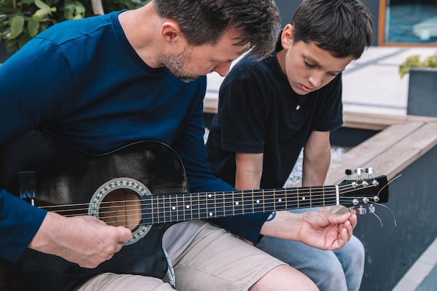 Zdjęcie młody ojciec uczy syna grać na gitarze letni dzień wolny na placu miejskim. koncepcja nowoczesnej szczęśliwej rodziny. styl życia.