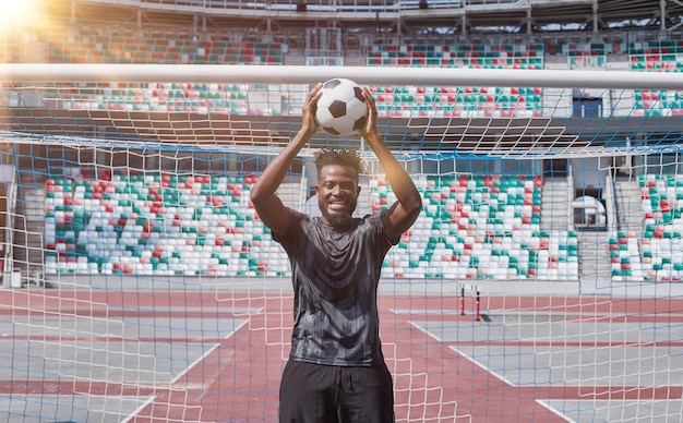 Młody, pewny siebie Afroamerykanin trzyma piłkę nożną w rękach na zewnątrz stadionu i patrzy w górę