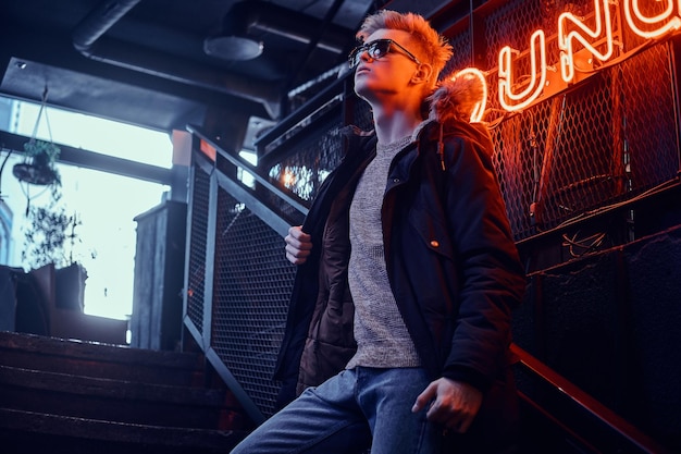 Zdjęcie młody pewny siebie facet w płaszczu z futrzanym kapturem i okularami przeciwsłonecznymi stojący na schodach do podziemnego klubu nocnego, w tle podświetlany szyld