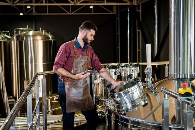 Młody piwowar mężczyzna w skórzanym fartuchu nadzorujący proces fermentacji piwa w nowoczesnym fa...