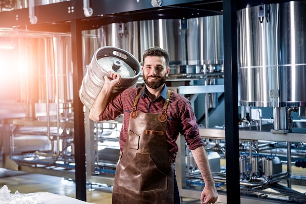Młody piwowar mężczyzna w skórzanym fartuchu trzyma beczkę z piwem rzemieślniczym w nowoczesnej fabryce browaru