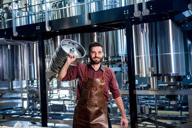Zdjęcie młody piwowar mężczyzna w skórzanym fartuchu trzyma beczkę z piwem rzemieślniczym w nowoczesnej fabryce browaru