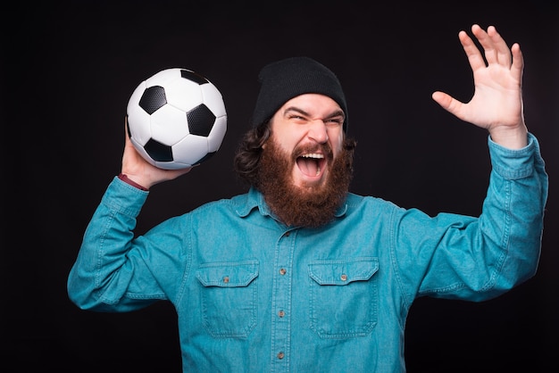 Młody podekscytowany brodaty mężczyzna krzyczy i trzyma piłkę w pobliżu czarnej ściany