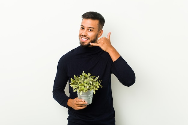 Młody południowoazjatycki mężczyzna trzyma rośliny pokazuje gesta telefonu komórkowego gesta palcami.