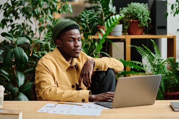 Młody poważny czarny człowiek w casual, siedzący przy miejscu pracy w zielonym biurze
