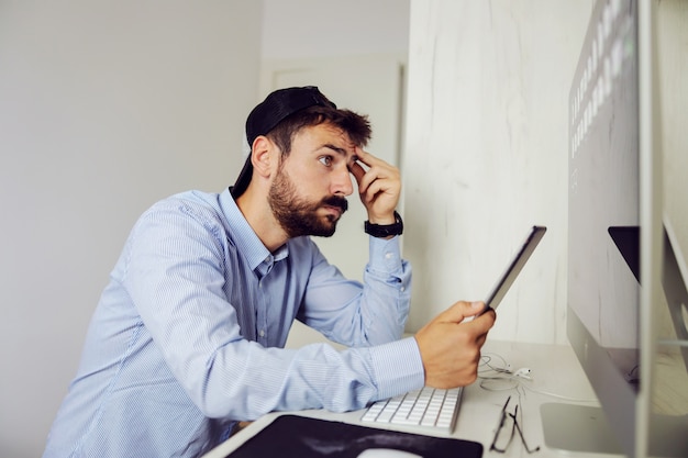 Młody poważny freelancer siedzi w domowym biurze, trzymając głowę i tablet podczas myślenia.