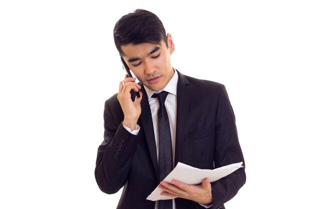 Młody pozytywny człowiek w białej koszuli i czarnym garniturze z krawatem, trzymając papiery i rozmawiający przez telefon