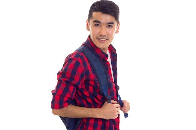 Młody pozytywny człowiek z czarnymi włosami w białej koszulce i czerwonej koszuli w kratkę z niebieskim plecakiem