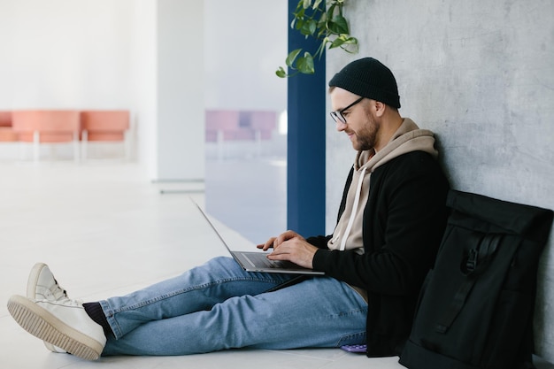 Młody programista używający laptopa piszący kod programowania siedząc na podłodze w nowoczesnym kreatywnym biurze startowym