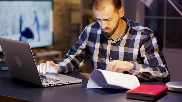 Młody przedsiębiorca pracuje na laptopie i podpisuje dokumenty w domowym biurze w godzinach nocnych.