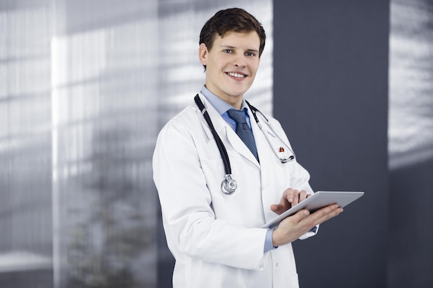 Młody przyjazny lekarz sprawdza niektóre informacje na swoim tablecie komputerowym Portret profesjonalnego lekarza przy pracy w klinice Pojęcie medycyny