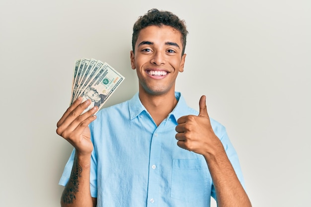 Młody przystojny afroamerykanin trzymający banknoty 20 dolarów uśmiechnięty szczęśliwy i pozytywny, kciuk w górę robi doskonały i znak zatwierdzenia