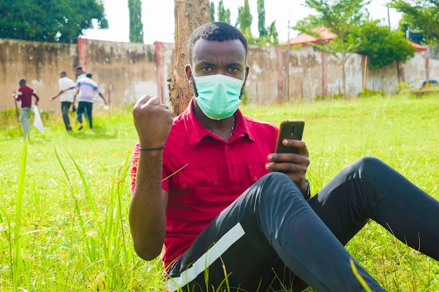 Zdjęcie młody przystojny afrykański mężczyzna siedzący na zewnątrz, noszący maskę na twarz, aby zapobiec, zapobiec, zapobiec wybuchowi epidemii w społeczeństwie podczas obsługi telefonu komórkowego