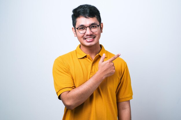 Młody przystojny azjatycki mężczyzna ubrany dorywczo koszula na białym tle z dużym uśmiechem na twarzy, wskazując palcem ręki w bok, patrząc w kamerę