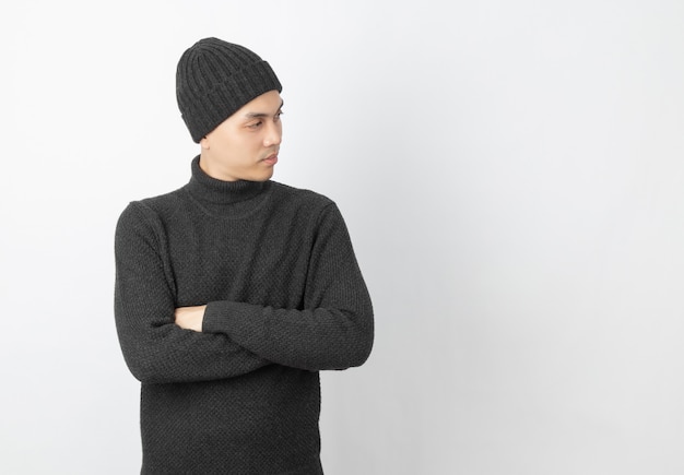 Młody przystojny azjatykci mężczyzna jest ubranym popielatego pulower i czapkę podczas gdy ręki krzyżować i przyglądający z szczęśliwą twarzą up na bielu