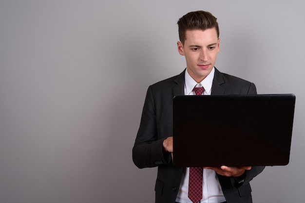 młody przystojny biznesmen ubrany w garnitur trzymając laptopa na szaro