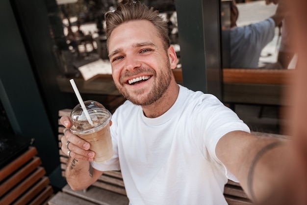 Młody przystojny blond białe zęby uśmiechnięty mężczyzna z zarost sprawia, że selfie z napojem filiżanka kawy nosi białą koszulkę na tarasie kawiarni z mobilnym smartfonem. Portret mężczyzny z bliska. Hipster milenijny