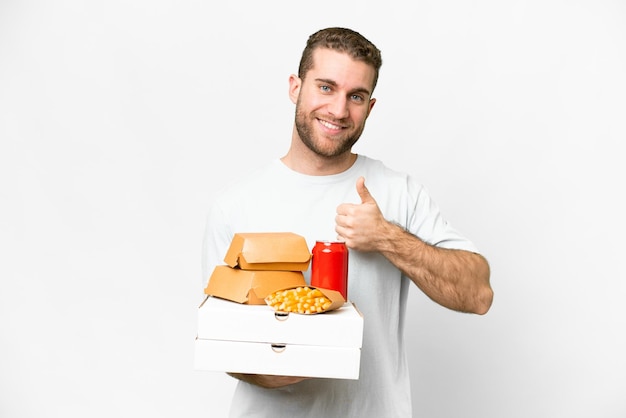 Młody przystojny blondyn trzymający pizze i hamburgery na odosobnionym tle, pokazujący gest kciuka w górę