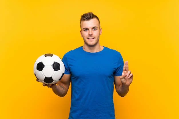 Młody przystojny blondynka mężczyzna trzyma piłki nożnej piłkę nad odosobnionym żółtym tłem wskazuje w górę doskonałego pomysłu
