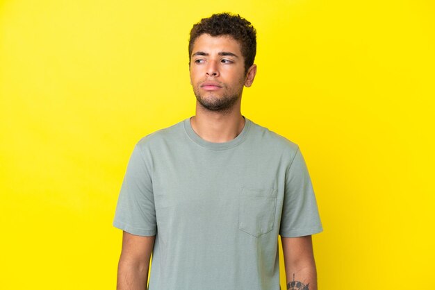 Młody przystojny brazylijski mężczyzna odizolowany na żółtym tle, mający wątpliwości, patrząc z boku