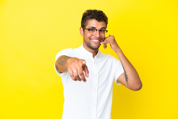 Młody przystojny brazylijski mężczyzna odizolowany na żółtym tle wykonujący gest telefonem i wskazujący przód