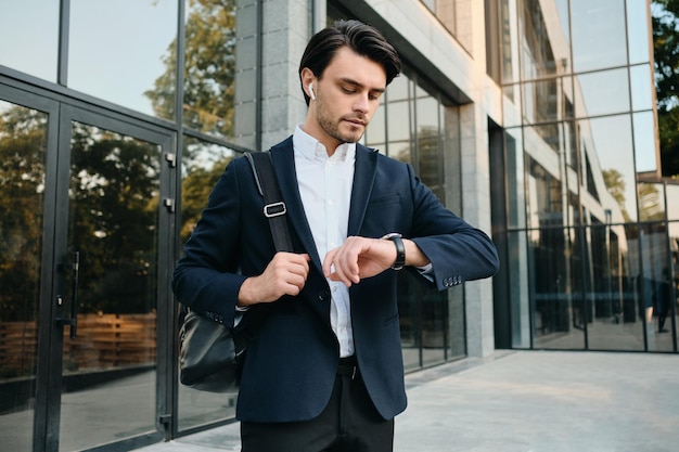 Młody przystojny brodaty mężczyzna brunetka w białej koszuli i klasycznym garniturze z bezprzewodowymi słuchawkami i plecakiem na ramieniu i marzycielsko patrząc na zegarek na zewnątrz
