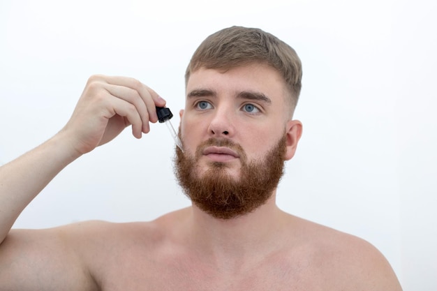 Młody przystojny brodaty mężczyzna stosuje balsam do produktu kosmetycznego z serum, aby uzyskać zdrowe, doskonałe, świeże nawilżenie skóry, zapuszczając brodę za pomocą nawilżającej pielęgnacji skóry na białym tle