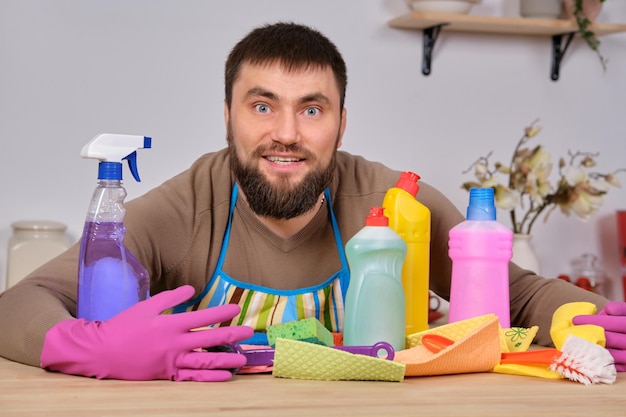 Młody przystojny brodaty mężczyzna w kuchni pokazuje cały swój personel sprzątający - detergenty, pędzle, spraye. Myśli, że jest gotowy na prawdziwe sprzątanie