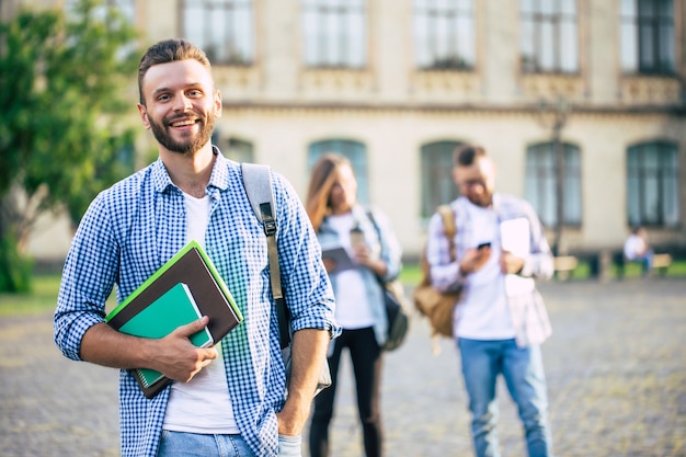 Młody przystojny brodaty student facet w ubranie z plecakiem i książkami w rękach chodzi i pozuje na budynkach uniwersytetu