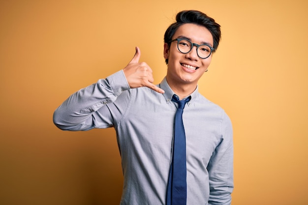 Młody przystojny chiński biznesmen w okularach i krawacie na żółtym tle uśmiechający się wykonujący gest telefoniczny ręką i palcami, jak rozmowa przez telefon