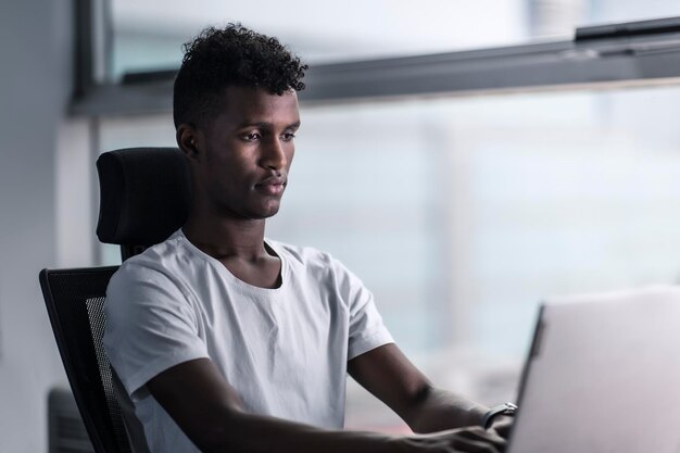 Młody przystojny czarny mężczyzna skoncentrowany na swoim laptopie i pracujący w poważnym nastroju w nowoczesnej przestrzeni biurowej