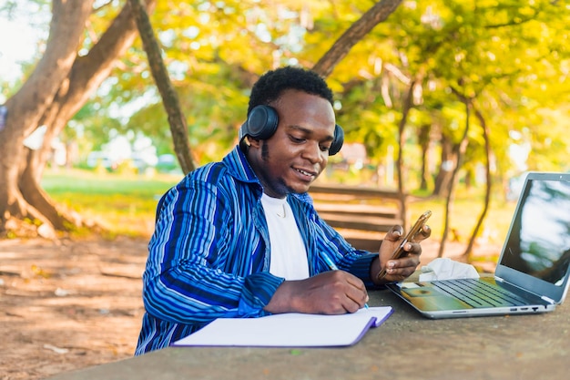 Młody przystojny czarny mężczyzna student siedzi pisania na papierze przy użyciu telefonu komórkowego w ręku