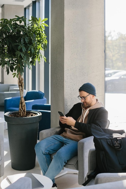 Młody przystojny facet z telefonem w dłoniach czeka na rozmowę kwalifikacyjną w biurze Mężczyzna w okularach i czapce trzyma telefon siedząc na krześle w holu