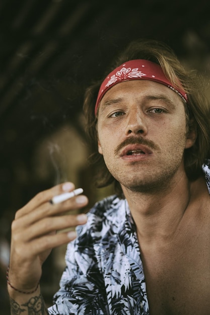 Młody przystojny hipis w czerwonej bandanie, z nagim torsem w kawiarni na Bali pali papierosa