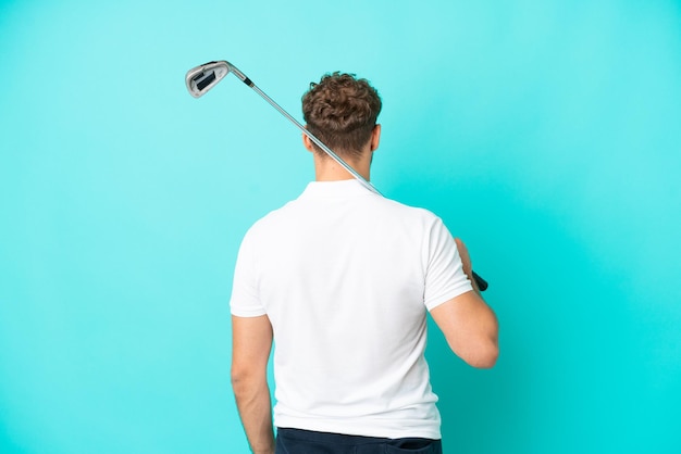 Młody przystojny kaukaski mężczyzna na białym tle na niebieskim tle, grając w golfa i w pozycji tylnej