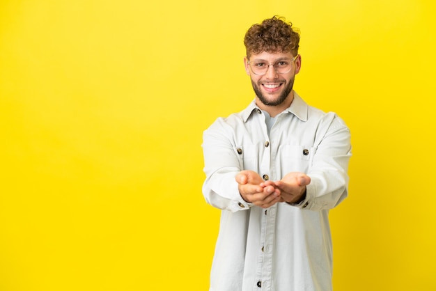 Młody przystojny kaukaski mężczyzna na żółtym tle trzyma wyimaginowaną copyspace na dłoni, aby wstawić reklamę