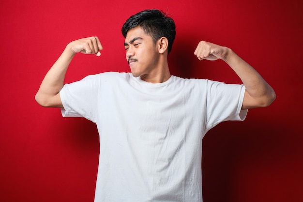 Młody przystojny mężczyzna azjatyckich na czerwonym tle na sobie biały tshirt pokazując mięśnie ramion uśmiechający się dumny. koncepcja fitness.