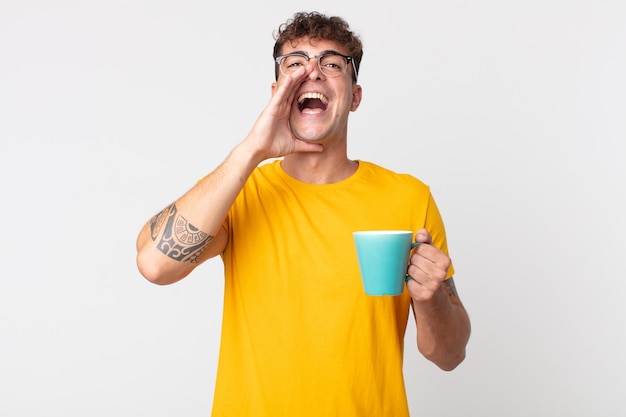 Młody przystojny mężczyzna czuje się szczęśliwy, wydając wielki okrzyk z rękami przy ustach i trzymając filiżankę kawy